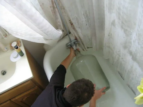 A person is filling a large glass container with water from a bathtub faucet, standing beside a shower curtain and sink with personal care items.