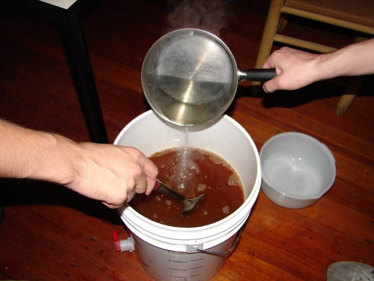 A person is pouring liquid from a kettle into a large white bucket with a spigot, as another person stirs the bucket's contents with a long spoon; a white bowl sits nearby on a wooden floor.