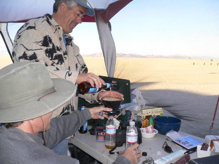 Two men at a campsite, one pouring a drink into a blender while the other watches, with a desert landscape in the background.