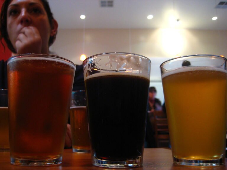 Three different types of beer in glasses on a table, with a woman pondering in the background.