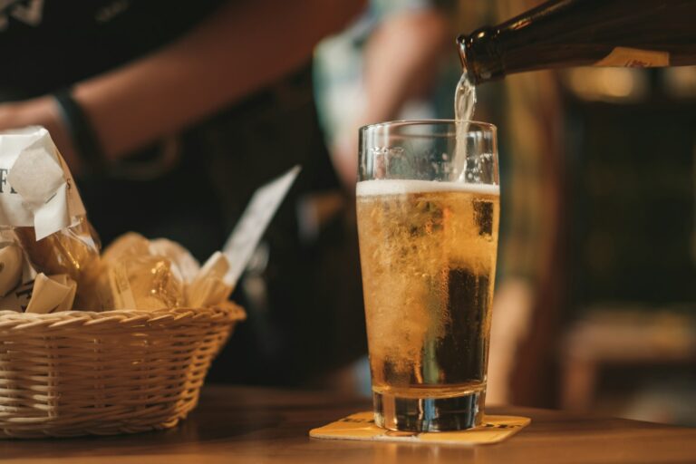 A beer bottle is being tilted to pour beer into a clear glass on a bar counter, with the glass partially filled and foam forming on top, beside a basket of packaged snacks.