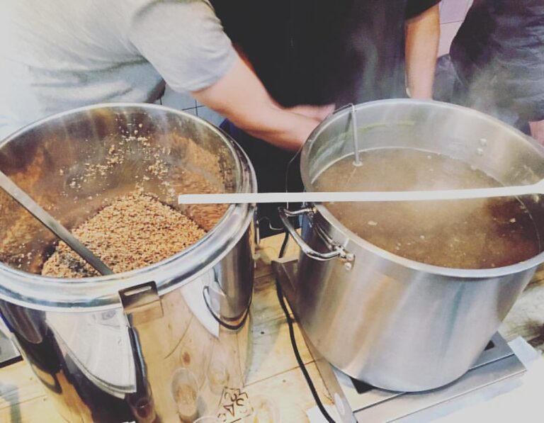Two large metal brewing vessels are shown, one with grains and one with liquid, possibly part of a beer brewing process, with steam rising from the hot liquid. Two individuals appear to be actively working with the equipment.