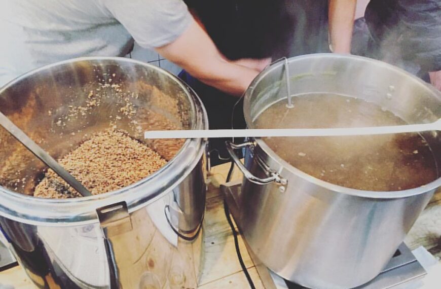 Two large metal brewing vessels are shown, one with grains and one with liquid, possibly part of a beer brewing process, with steam rising from the hot liquid. Two individuals appear to be actively working with the equipment.