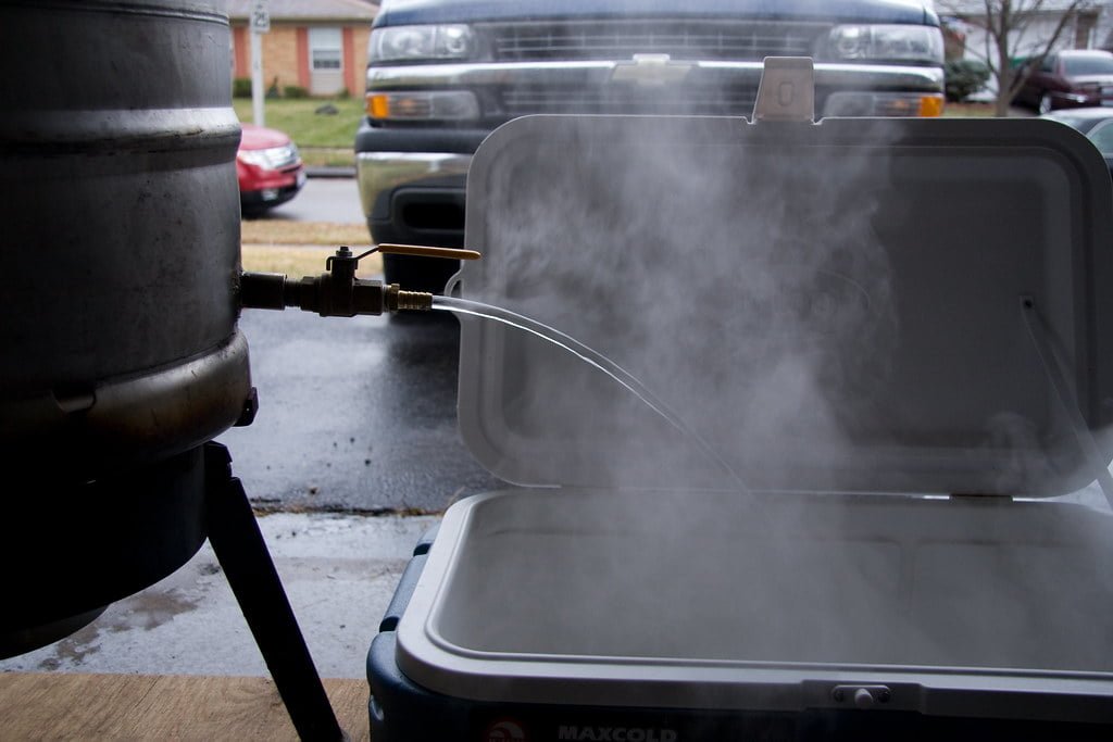 A large pot connected to a propane tank with steam flowing from a tap into an open cooler in an outdoor setting, with parked cars in the background.