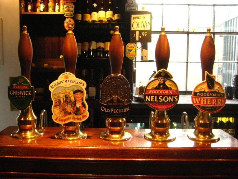 Five traditional beer pumps on a bar with labeled handles for Fuller's Chiswick Bitter, Batemans Bloomin' Marvellous, Theakston Old Peculier, Woodforde's Nelson's, and Woodforde's Wherry. A window and shelves with bottles and glasses are visible in the background.