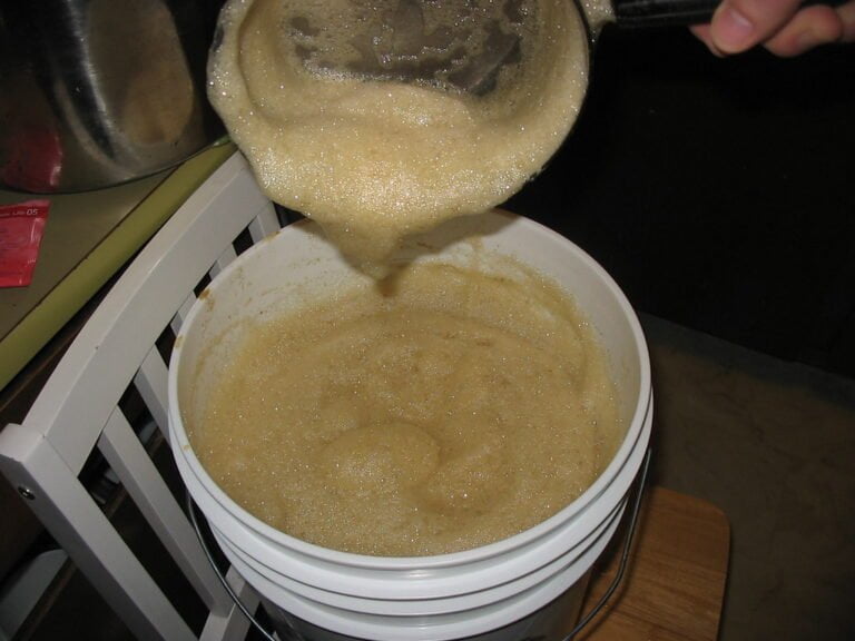 A person is pouring a foamy liquid from a brewing kettle into a fermentation bucket during a homebrewing process.