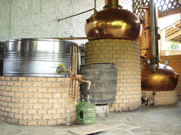 A distillery setup with copper pot stills, brick bases, and stainless steel tanks in a room with white walls and stone flooring.