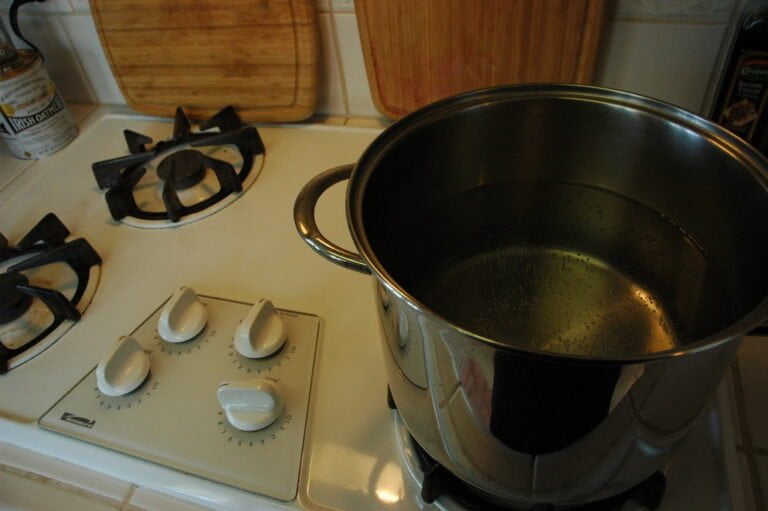 A stainless steel pot with water heating on a white gas stove top with burner dials in the foreground.