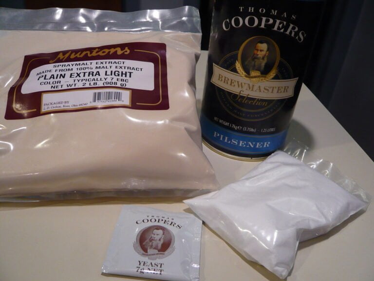 Homebrew beer ingredients on a table, including a package of Muntons Spraymalt Extract, a can of Thomas Coopers Brewmaster Selection Pilsner malt concentrate, and a packet of Thomas Coopers brewer's yeast.