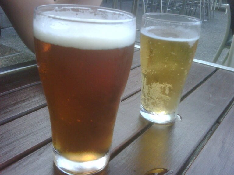 Two glasses of beer on a wooden table, one with amber-colored beer and a frothy head, the other with pale yellow beer and less foam.