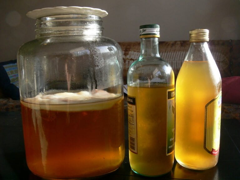 A large glass jar with a white substance on top contains a translucent amber liquid, flanked by two bottles filled with similar colored liquid, all illuminated by sunlight.