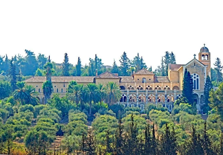 A historic stone-built monastery with arched windows surrounded by a lush garden of palm trees and shrubs under a clear sky.