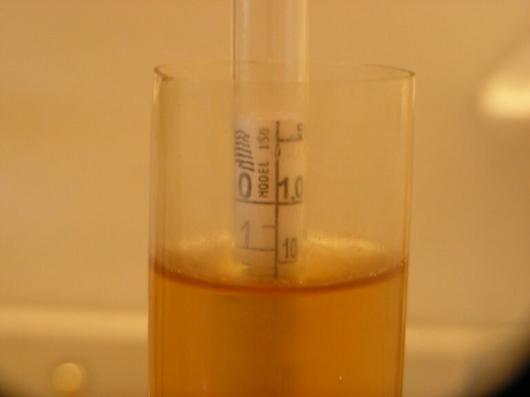 A close-up of a hydrometer floating in a cylinder containing amber liquid, with the scale on the hydrometer in focus showing specific gravity levels.