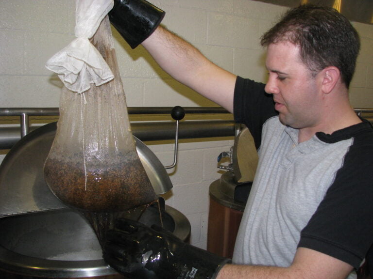A person wearing a black glove holding a large bag of grain above a stainless-steel brewing kettle, allowing liquid to drain from the bag into the kettle.