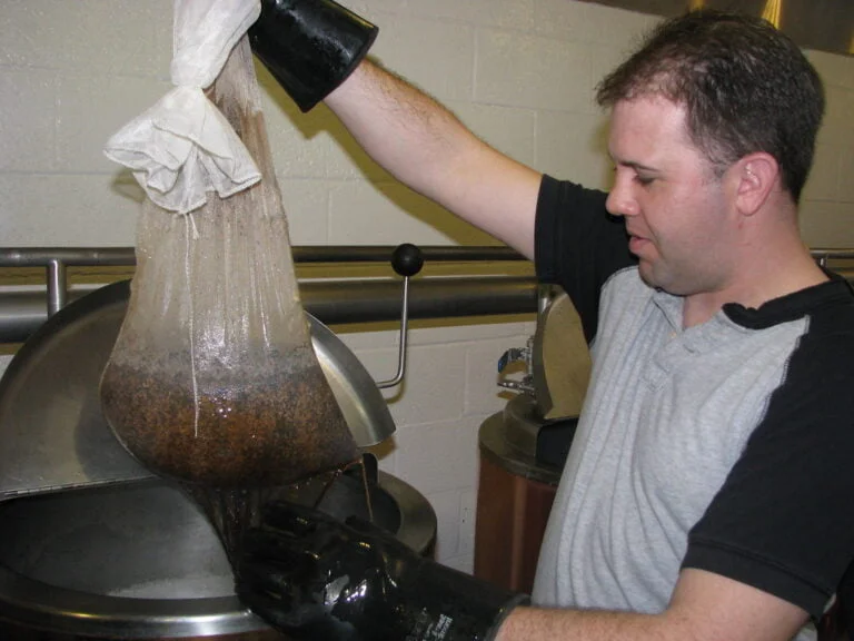 A person wearing a black glove holding a large bag of grain above a stainless-steel brewing kettle, allowing liquid to drain from the bag into the kettle.