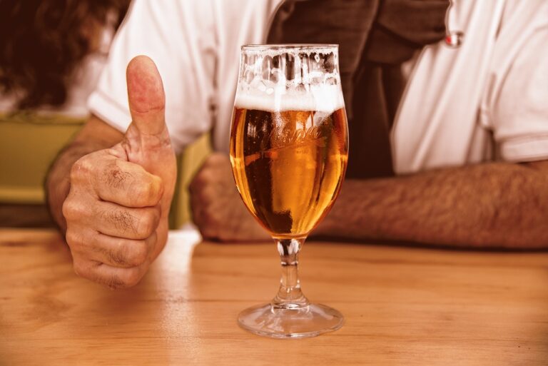 A person giving a thumbs-up sign next to a half-full glass of beer on a wooden table.