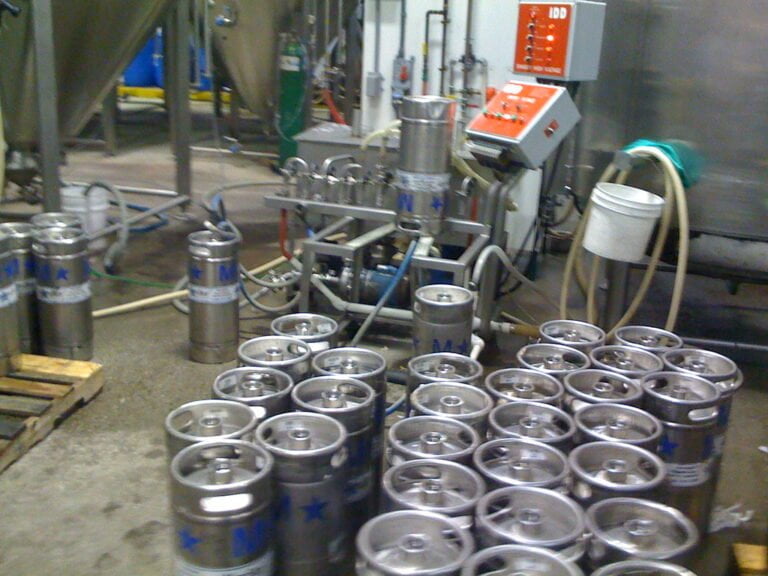 A brewery floor with multiple stainless steel beer kegs, some upright and some lying down, with hoses and brewing equipment in the background.