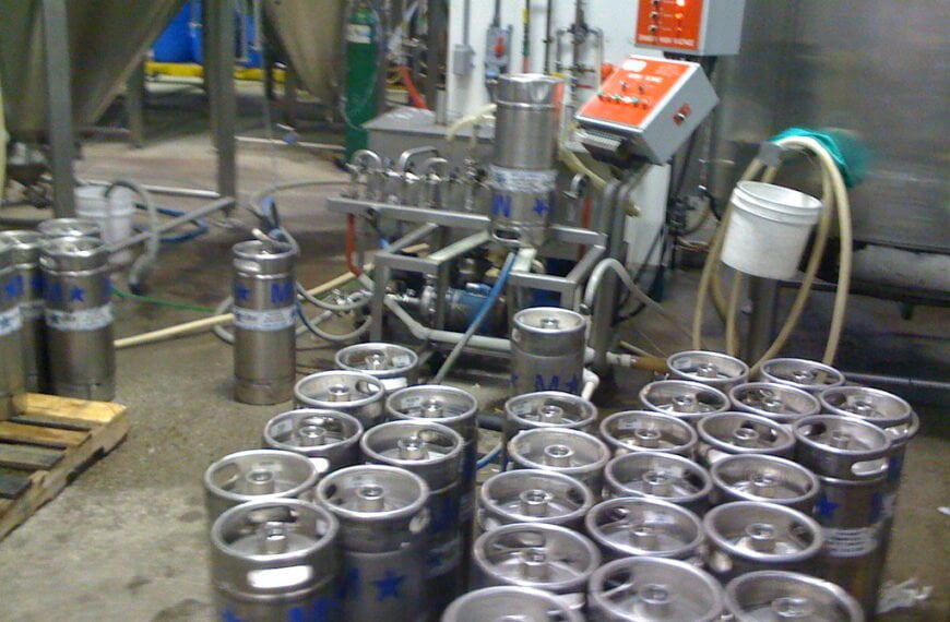 A brewery floor with multiple stainless steel beer kegs, some upright and some lying down, with hoses and brewing equipment in the background.