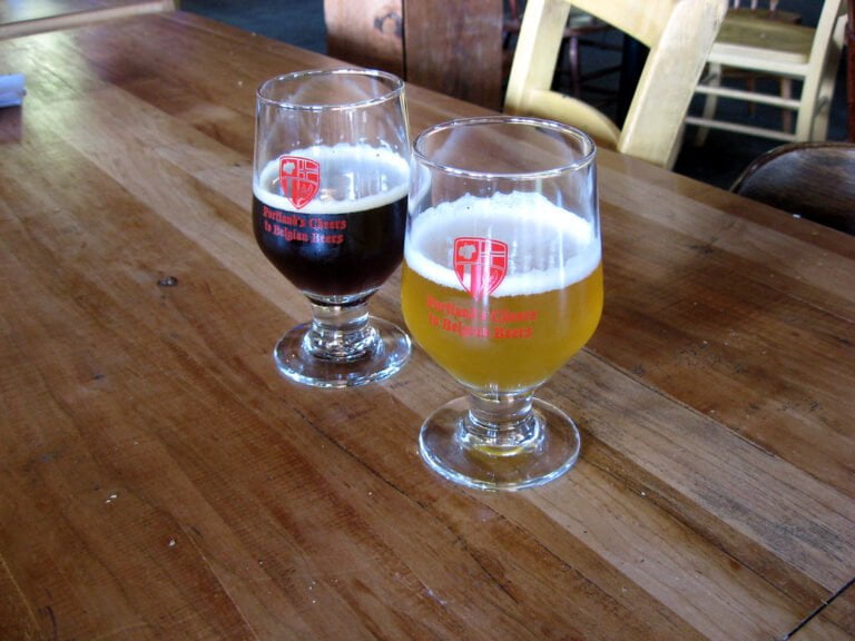 Two glasses of beer with logos reading "Portland's Cheers to Belgian Beers" on a wooden table, one glass half full of dark beer and the other mostly full of pale beer.