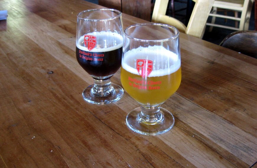 Two glasses of beer with logos reading "Portland's Cheers to Belgian Beers" on a wooden table, one glass half full of dark beer and the other mostly full of pale beer.