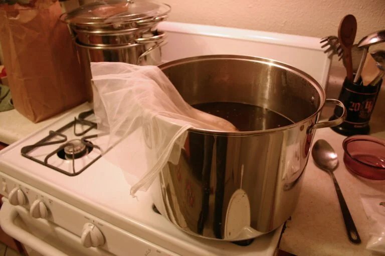 A large stainless steel pot on a white gas stove with a cheesecloth hanging over the side, surrounded by various cooking utensils and a paper bag.
