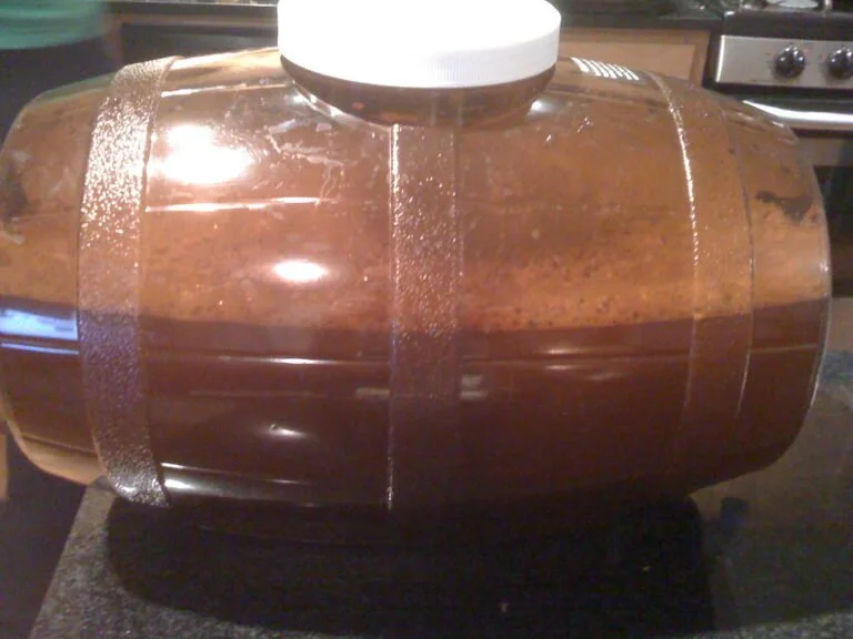 A large plastic jug filled with amber liquid, possibly homemade beer or cider, with sediment settled at the bottom, placed on a kitchen countertop.