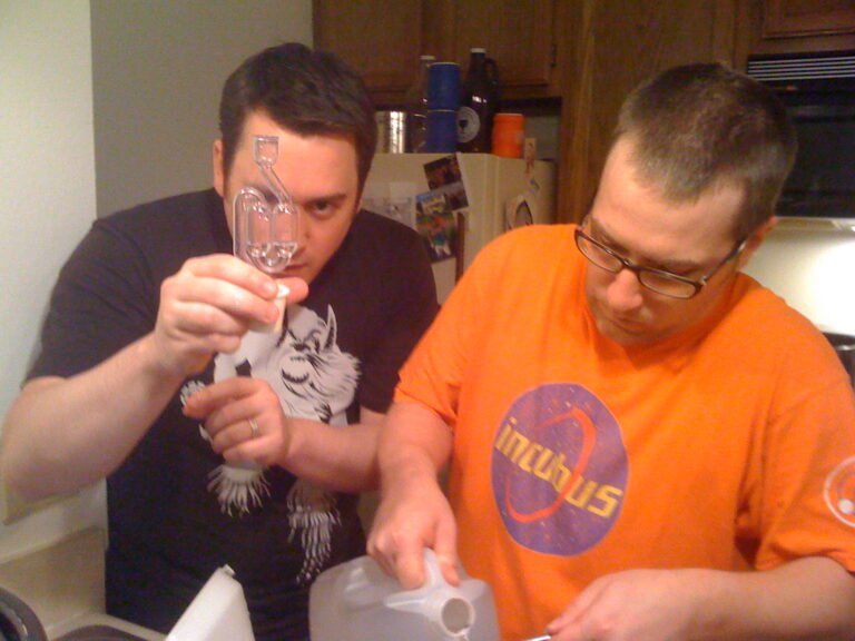 Two men in a kitchen, one pouring milk into a glass while the other holds the glass and looks at it intently.