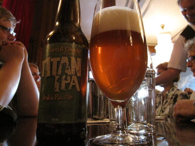 A bottle of Great Divide Titan IPA is placed on a tabletop next to a full glass of amber-colored beer with a frothy head. In the background, blurred figures of people appear to be engaged in conversation.