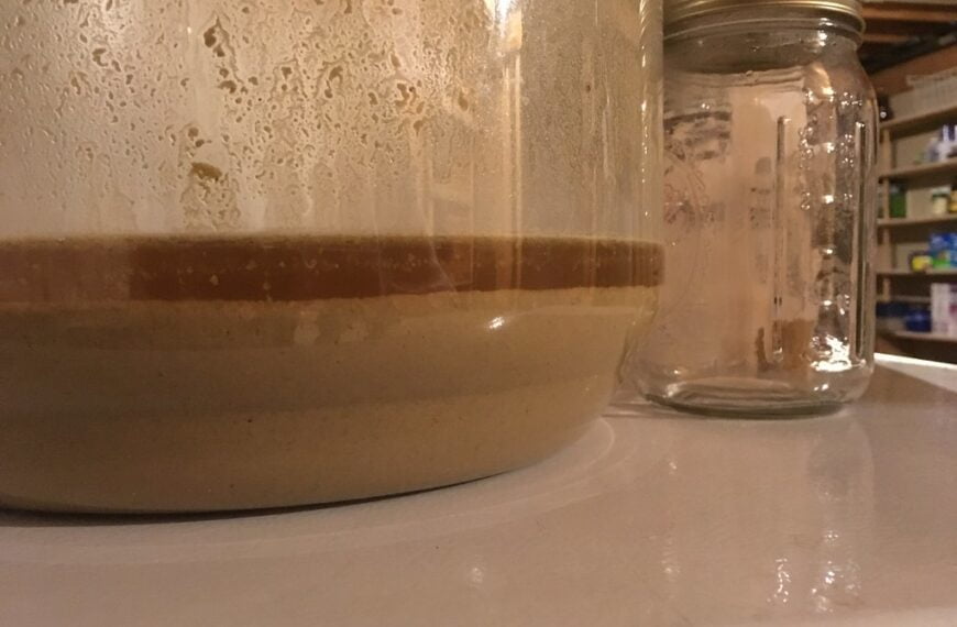 A close-up image showing two glass jars on a white surface, one large jar with visible layers of a brown sediment and a thicker beige substance on top and a smaller empty mason jar with a metal lid to the right. Droplets of condensation are visible on the inside surfaces of both jars.
