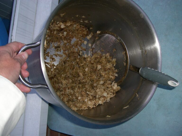 A person's hand holding a metal sieve over a stainless steel pot filled with what appears to be cooked, chopped food on a blue countertop.