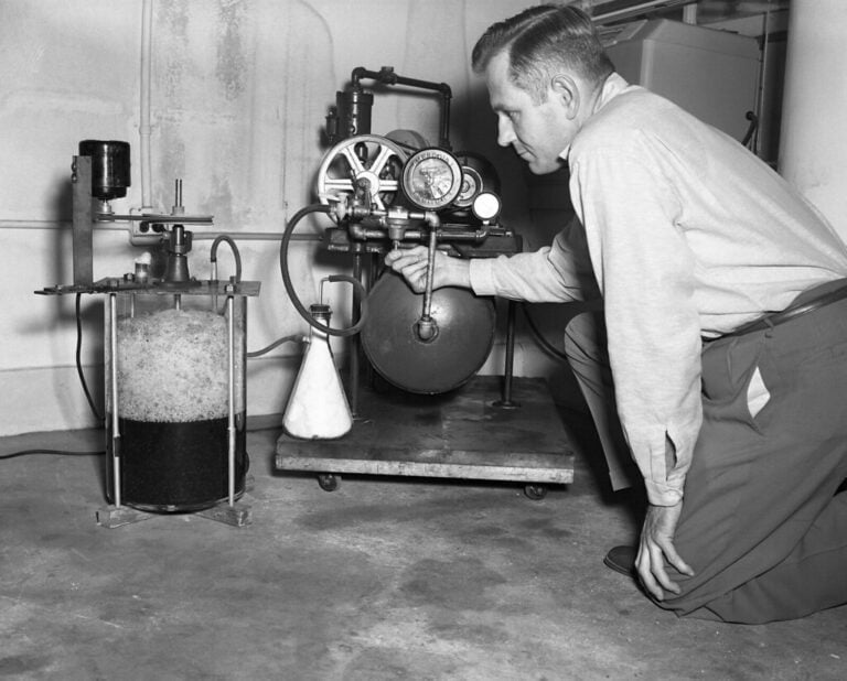 A black and white photo showing a man in a long sleeve shirt and trousers kneeling beside scientific equipment, including a pressure gauge and a glass container filled with a bubbly liquid.