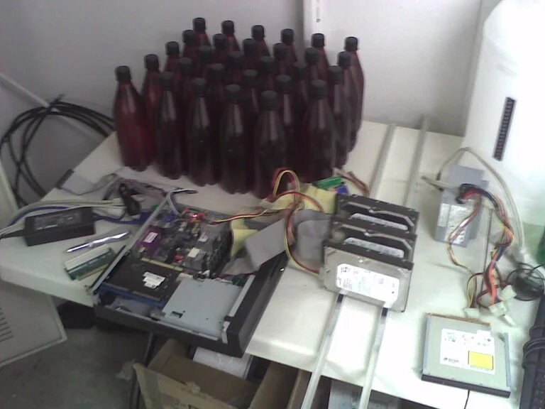 A cluttered workspace with an open computer case, internal components and cables on a table alongside rows of brown bottles and a large white container.