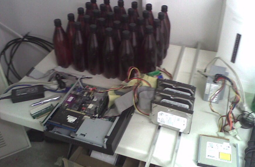 A cluttered workspace with an open computer case, internal components and cables on a table alongside rows of brown bottles and a large white container.