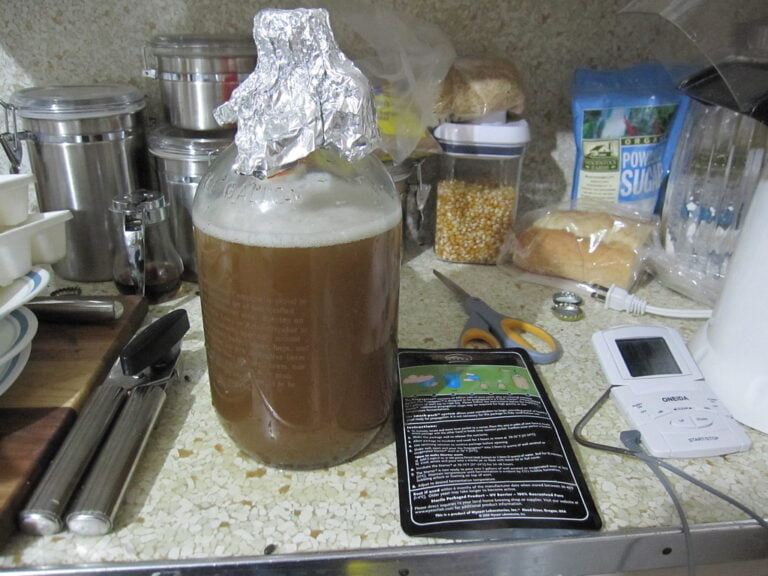 A cluttered kitchen countertop with various items including a bottle covered with aluminum foil overflowing with foam, a smartphone, a blood pressure monitor, kitchen utensils, and scattered food items like popcorn kernels and powdered sugar.