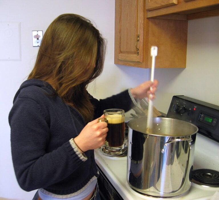 A person stirring a large pot on a stove while holding a mug of dark liquid in the other hand.