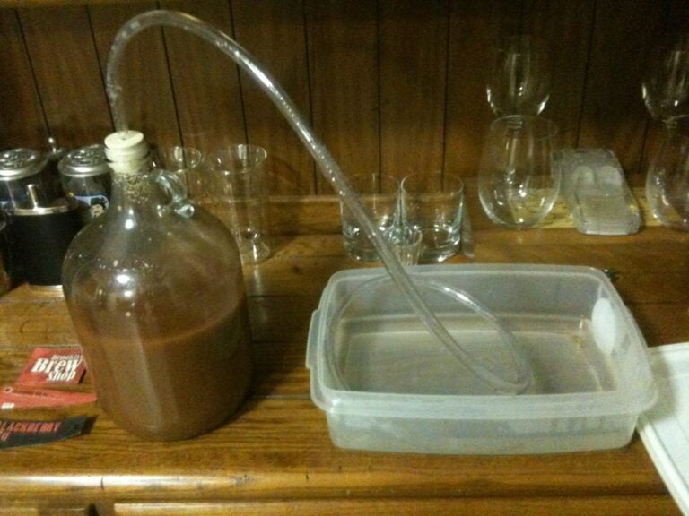 A glass carboy filled with a brown liquid connected via a siphon hose to a plastic container on a wooden surface, with glassware in the background and a homebrew book visible to the left.