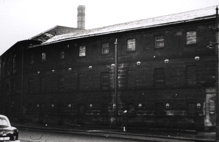 A black and white photograph of a large, old industrial building with multiple small windows and a single chimney stack, under a light dusting of snow. A vintage car is partially visible in the foreground on the left.