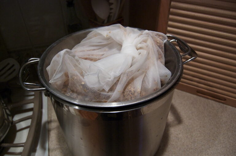 A stainless steel pot on a kitchen counter with a white cheesecloth full of ingredients being used to make stock.
