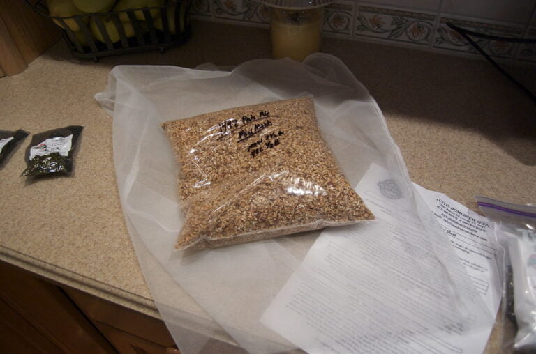 A large clear plastic bag of grain sits on a kitchen countertop next to a smaller packet of hops, a sheet of paper with text, and some other obscured kitchen items. A bowl of apples and a jar is partially visible in the background.