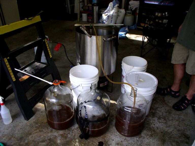 Home brewing setup in a garage with fermentation in glass carboys, a large metal brewing kettle, and various brewing equipment. A person in shorts is partially seen standing to the right.