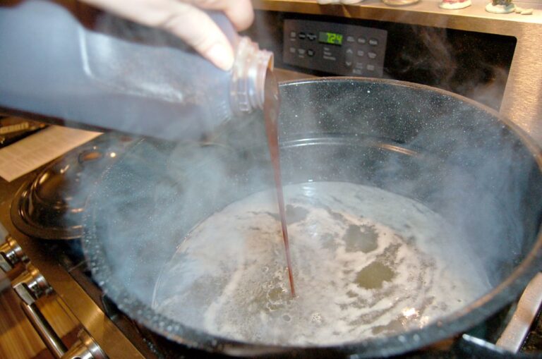 A person is pouring a liquid from a plastic bottle into a steaming pot on a stove, with a digital clock reading "12:41" in the background.