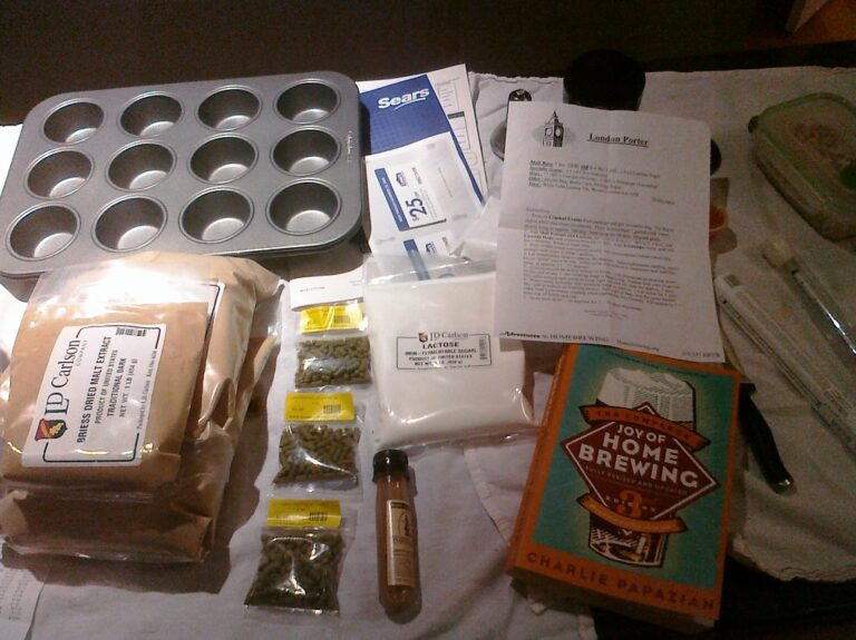 Homebrewing ingredients and equipment laid out on a table, including malt extract, hops, yeast, a brewing guide book titled "The Complete Joy of Home Brewing," and a recipe sheet for London Porter. A muffin tin and a hydrometer are also visible.