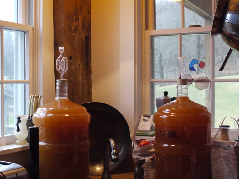 Two large carboys filled with liquid, one with an airlock, the other with a blow-off tube, positioned on a kitchen counter next to a window.