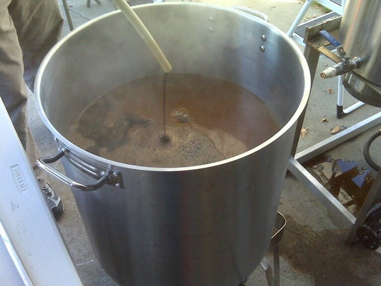 A large metal brewing kettle filled with liquid is being stirred with a long plastic paddle. The kettle is supported by a stand over a concrete floor, and there is brewing equipment visible in the background.