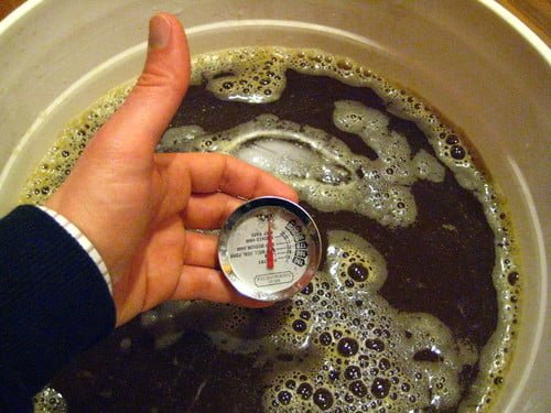 A person's hand holding a thermometer submerged in a bucket of dark liquid with foam on the surface.