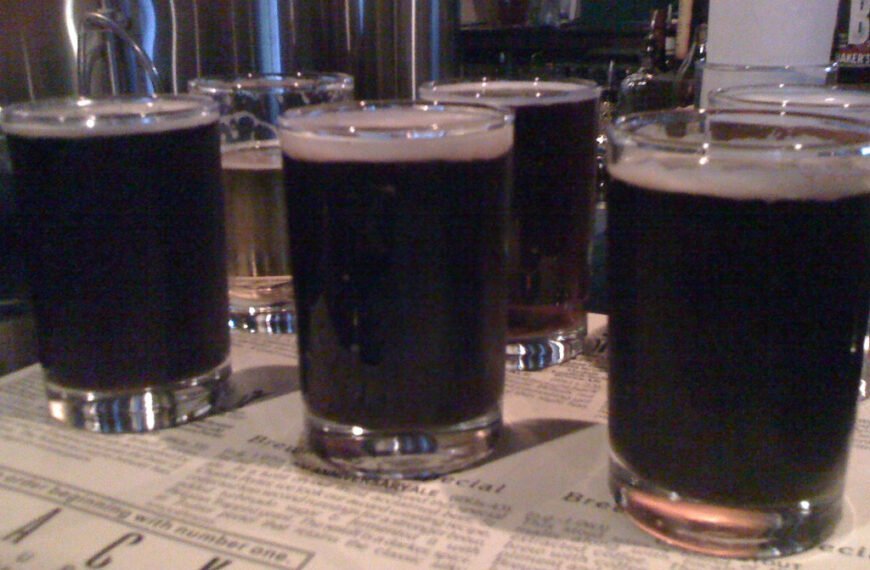 Four glasses of dark beer on a bar counter with a menu in the foreground and taps in the background.
