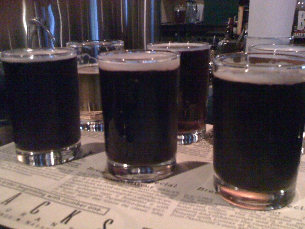 Four glasses of dark beer on a bar counter with a menu in the foreground and taps in the background.