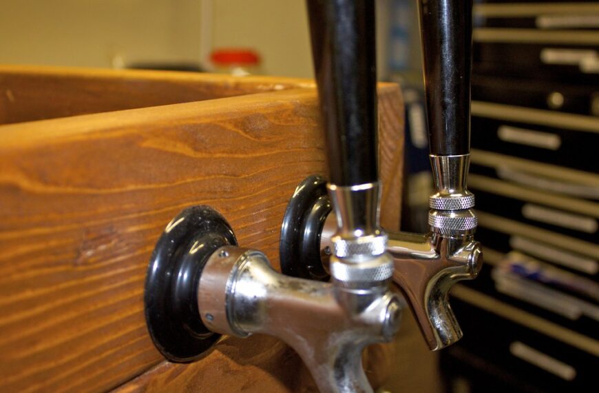 Two beer tap handles mounted on a wooden bar with a blurry background featuring workshop tools.