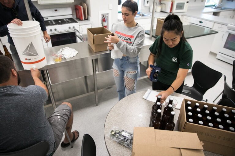 Three people are engaged in a brewing process with bottles and equipment on a kitchen island. On the left, a person is holding a white bucket with a spigot near a small glass, suggesting they might be transferring liquid. In the center and right, two people are interacting with bottles; one is holding a camera and the other seems to be inspecting or labeling the bottles. There are boxes and kitchen appliances in the background.