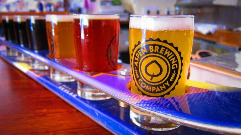 A flight of various beers lined up on a bar with a clear glass in the foreground featuring the Aspen Brewing Company logo.